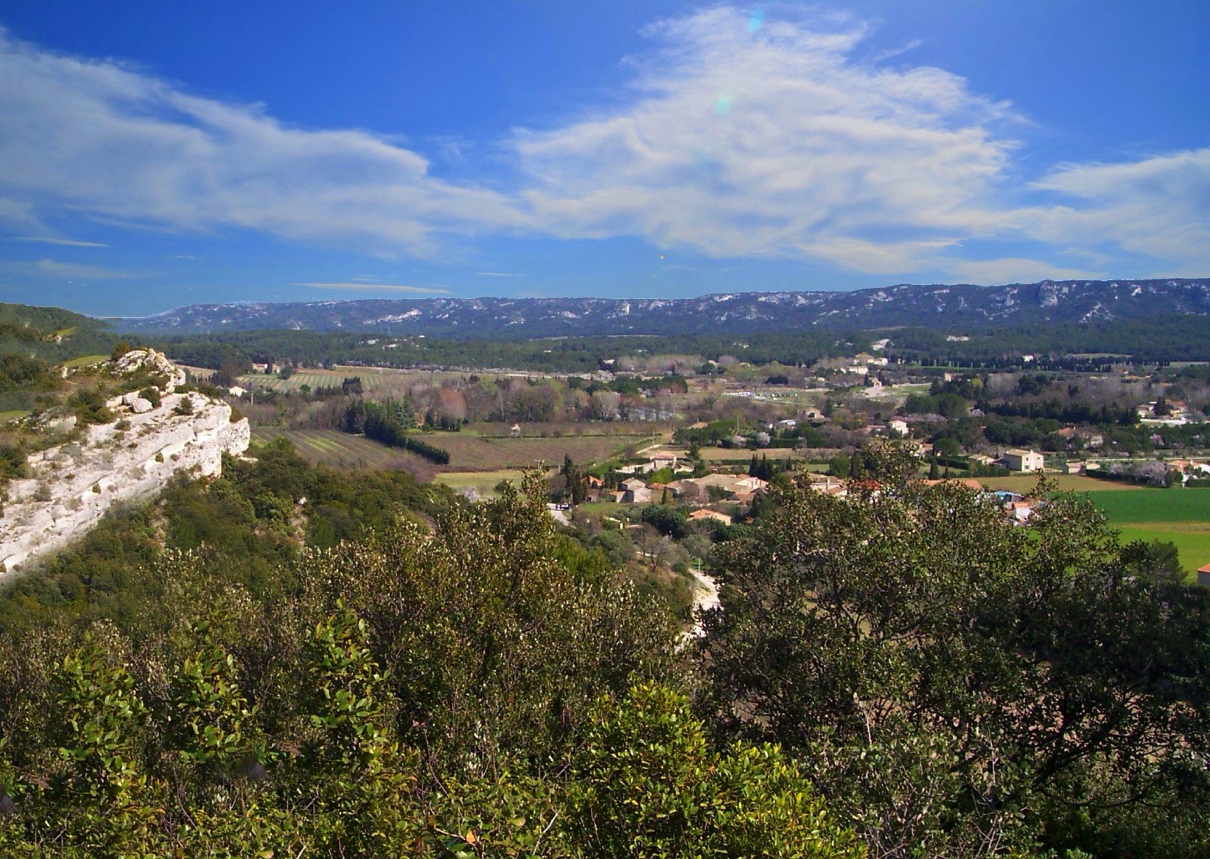 Panoramic view of a lush valley with scattered buildings, surrounded by rolling hills and rocky outcrops under a clear blue sky.